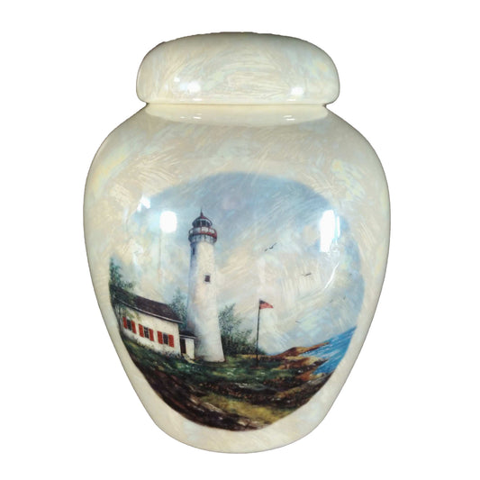 A beautiful small-sized cremation ceramic keepsake urn with a decoration of a lighthouse on a rocky shorline with a USA flag on the flagpole with a mother-of-pearl finish.