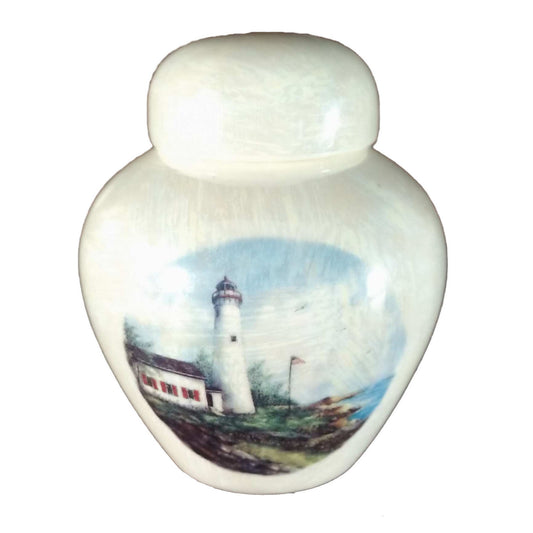 A beautiful small sized cremation ceramic keepsake urn with a decoration of a lighthouse on a rocky shoreline with a USA flag on the flagpole with a mother-of-pearl finish.