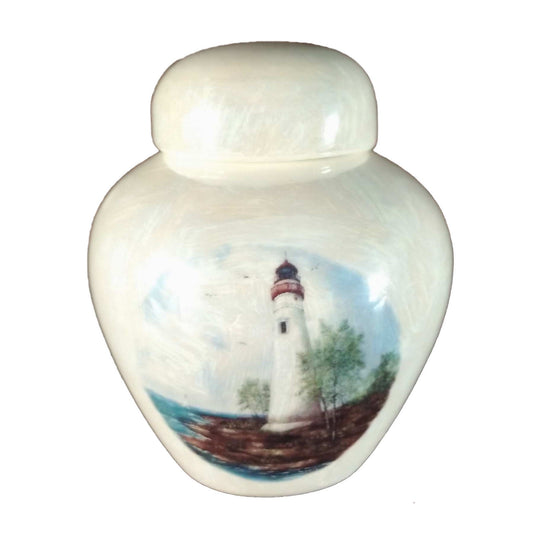 A beautiful keepsake cremation ceramic keepsake urn with a decoration of a lighthouse sitting on a rocky shoreline with a mother-of-pearl finish.